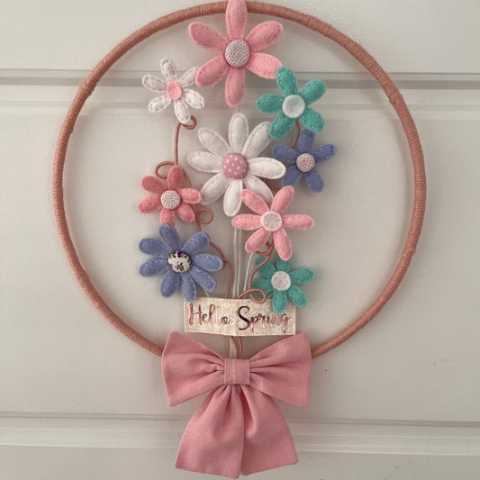 A round, yarn-wrapped wreath with felt flowers, a Hello Spring sign, and a pink bow.