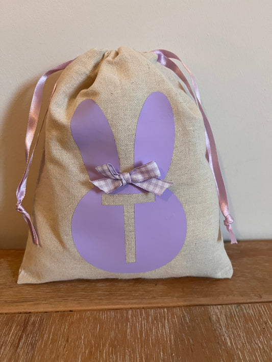 A beige drawstring bag with a purple bunny silhouette and a plaid bow, featuring a T cutout.