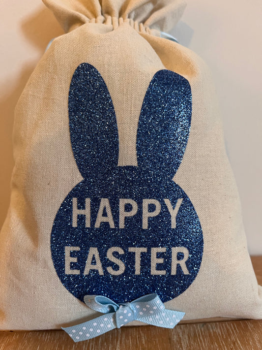 A beige drawstring bag with a blue glitter bunny silhouette and Happy Easter text.