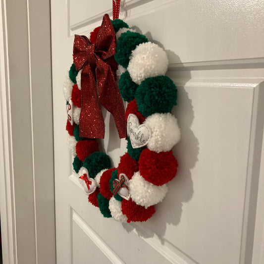 Handmade Christmas wreath with red, green, and white pom-poms, a red bow, and small white ornaments.
