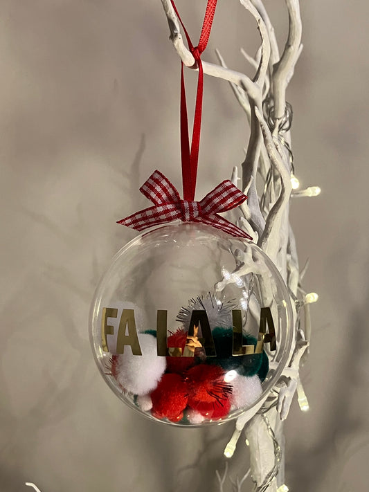 Clear Christmas ornament with "FA LA LA" text, red and white pom-poms, and a plaid bow.