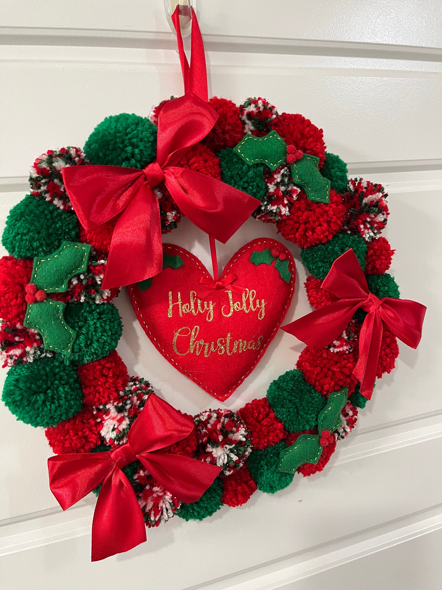 A handmade Christmas wreath with red and green pompoms, bows, and a 'Holly Jolly Christmas' heart.