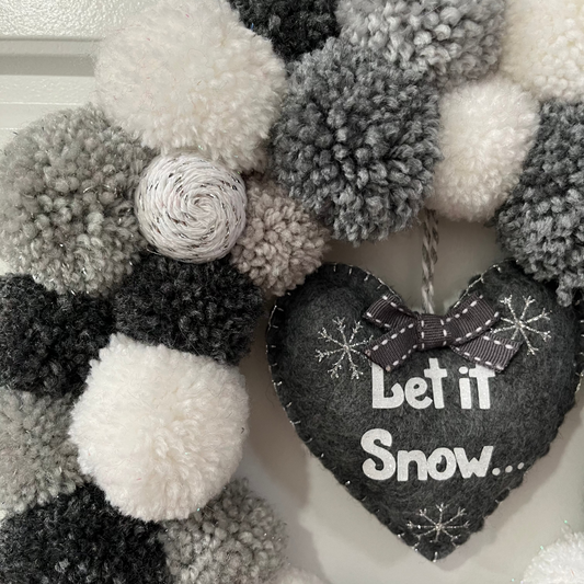 Close-up of a festive wreath with gray, black, and white pompoms and a “Let it Snow” heart.