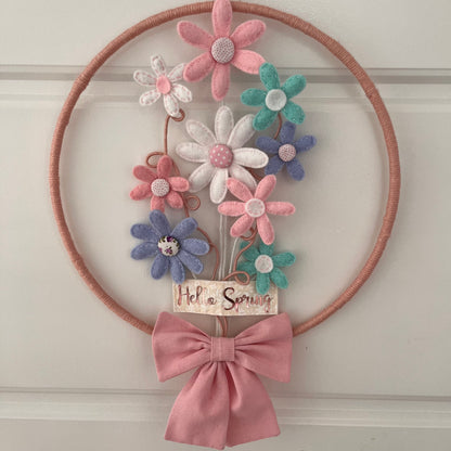 A round, yarn-wrapped wreath with felt flowers, a Hello Spring sign, and a pink bow.
