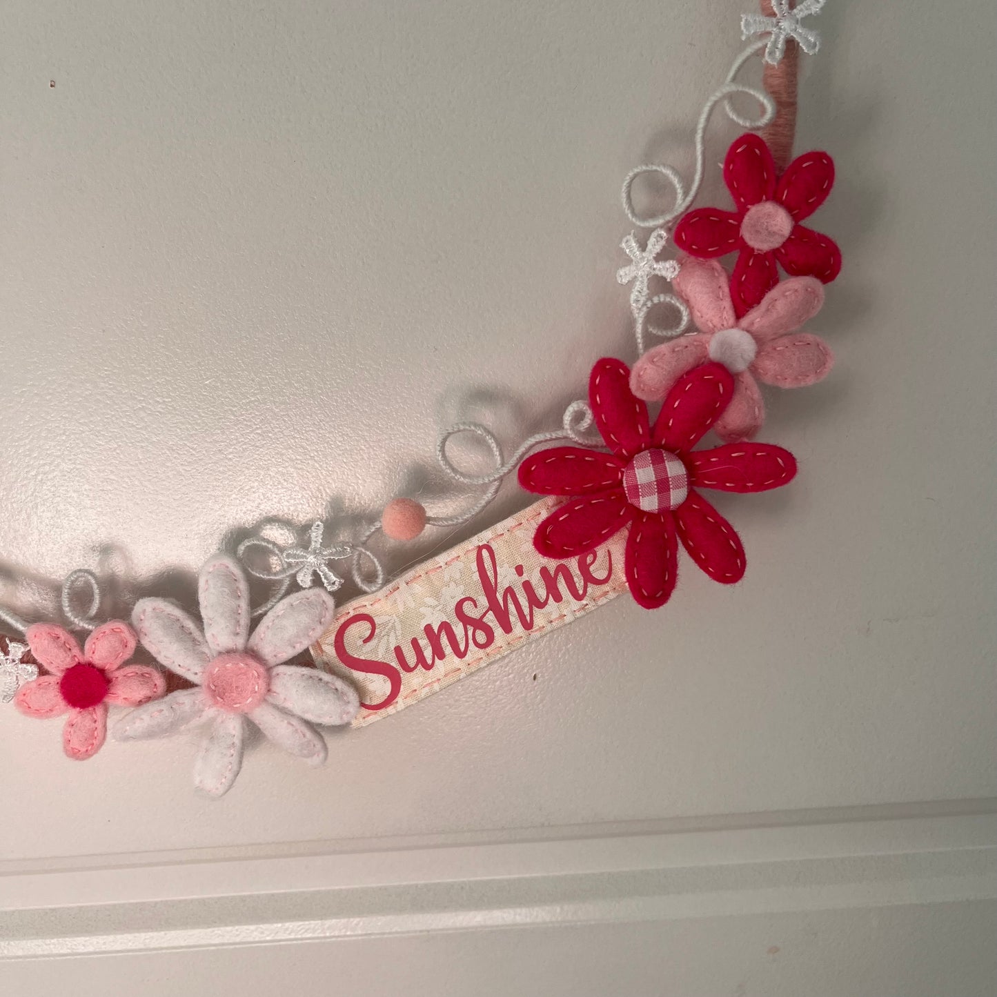 Close-up of a decorative wreath with felt flowers in shades of pink and red, and a ribbon reading Sunshine.