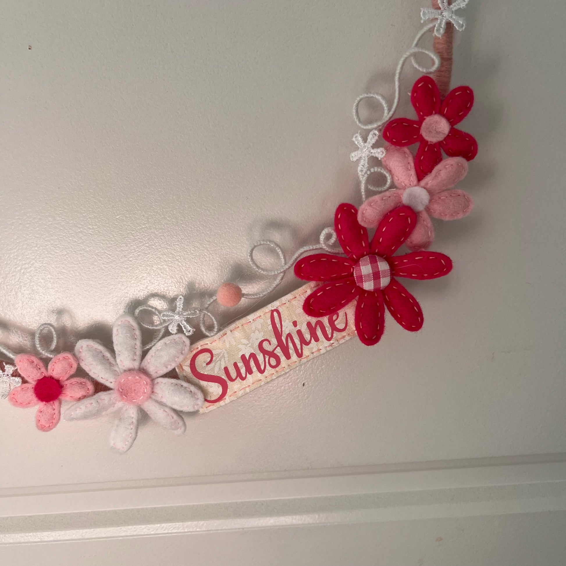 Close-up of a decorative wreath with felt flowers in shades of pink and red, and a ribbon reading Sunshine.