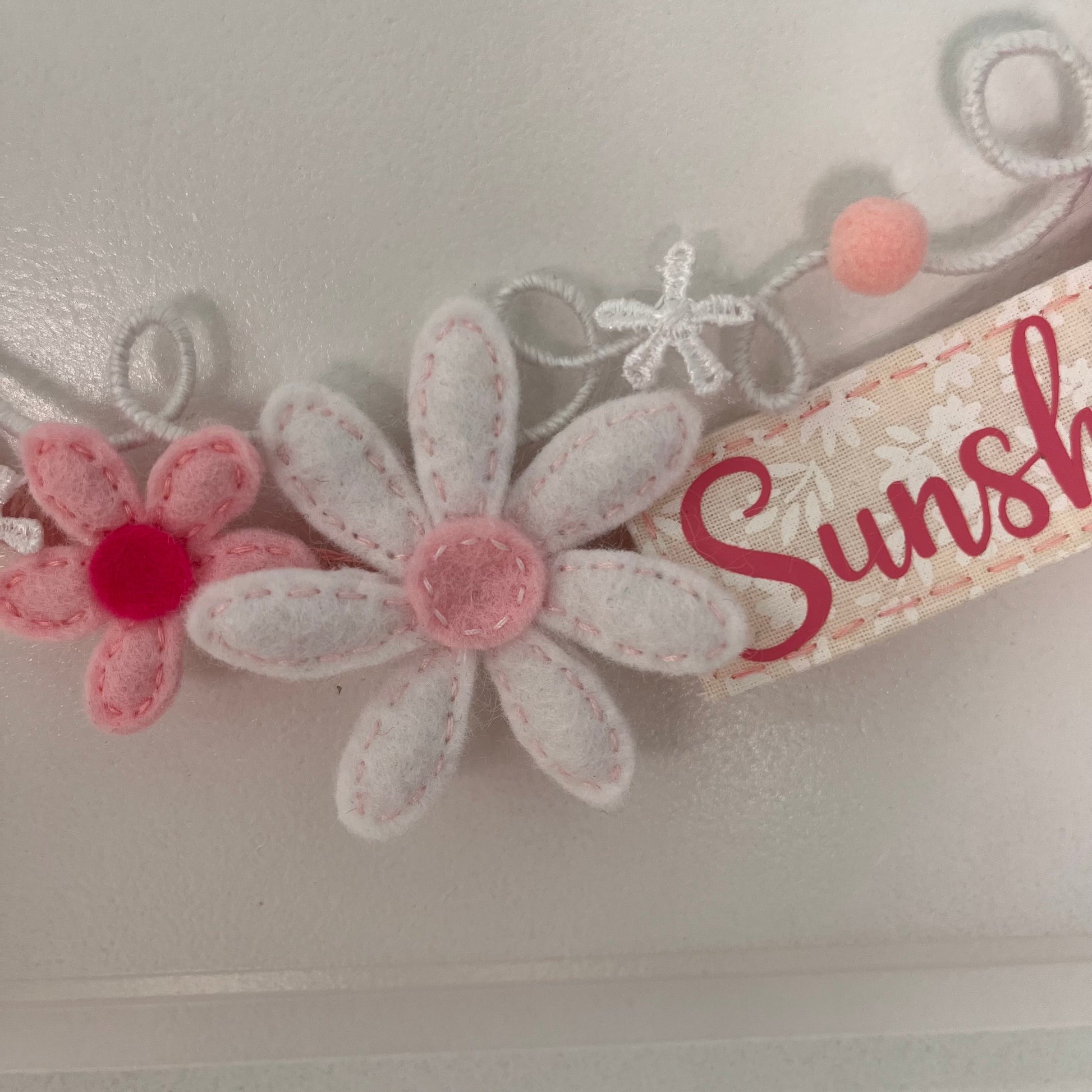 Close-up of a decorative item featuring felt flowers in pink and white, with a ribbon displaying part of the word Sunshine.