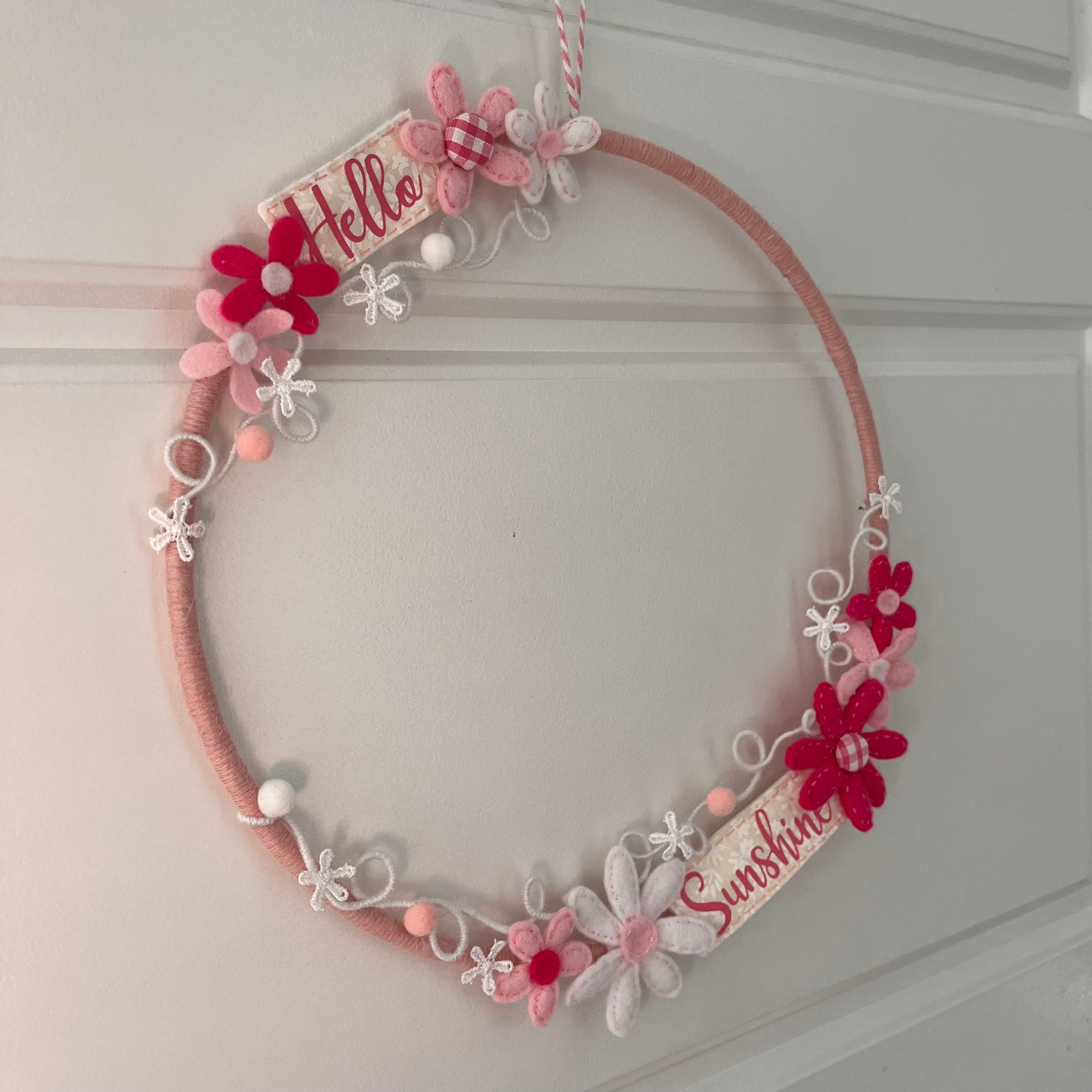 Pink yarn-wrapped wreath with felt flowers and Hello Sunshine signs, hanging on a white door.