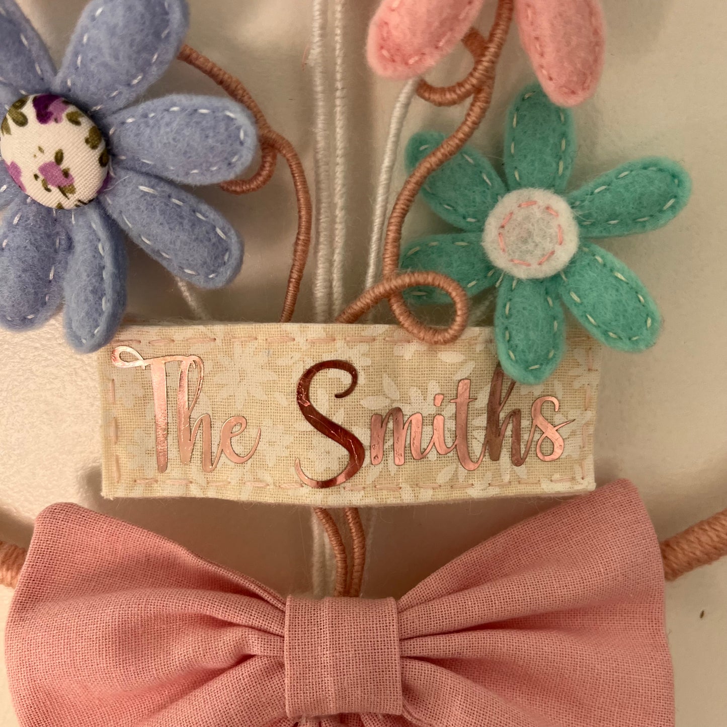 Close-up of a decorative wall hanging with felt flowers, a pink bow, and a fabric tag reading The Smiths.