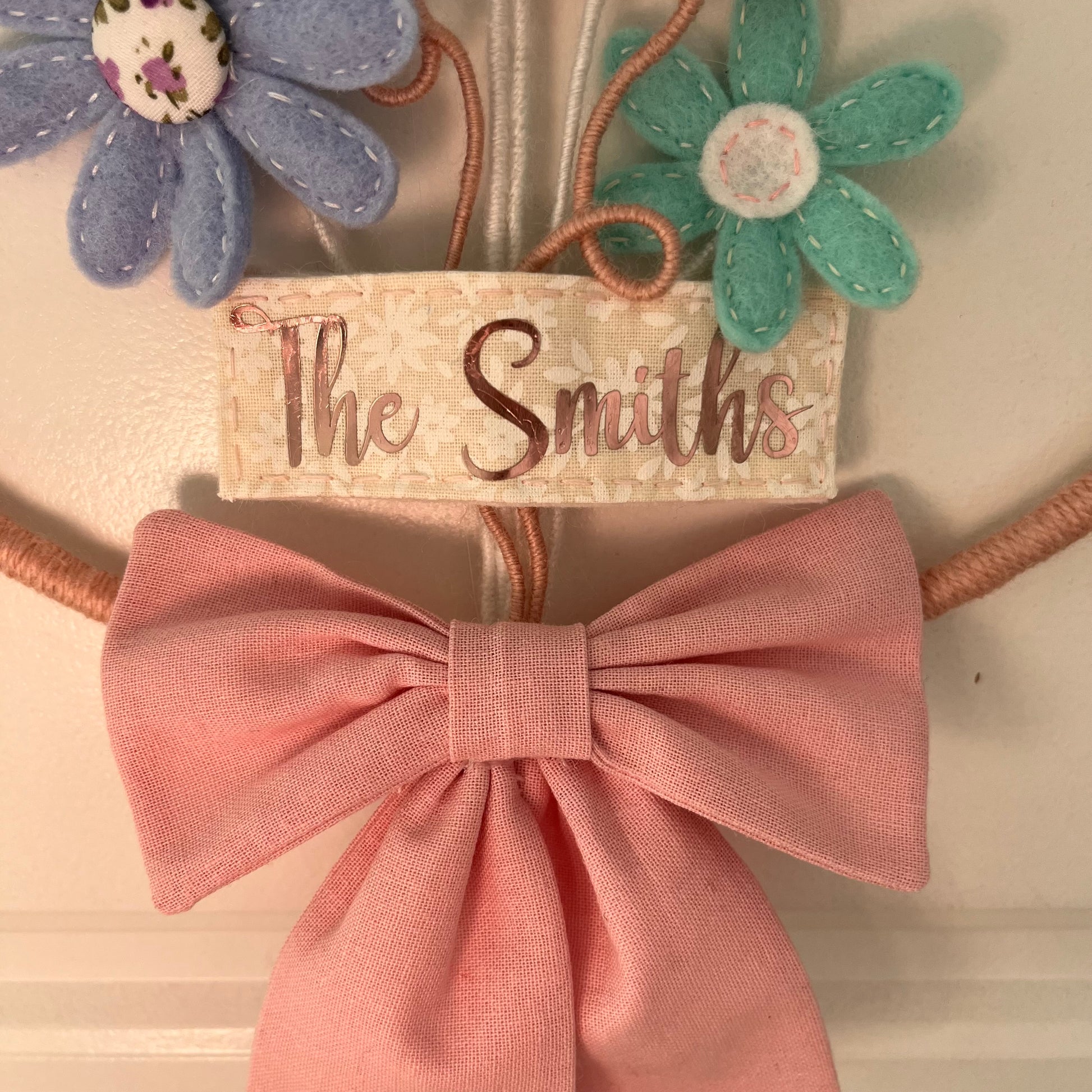 Close-up of a decorative wall hanging with felt flowers, a fabric tag reading The Smiths, and a large pink bow.