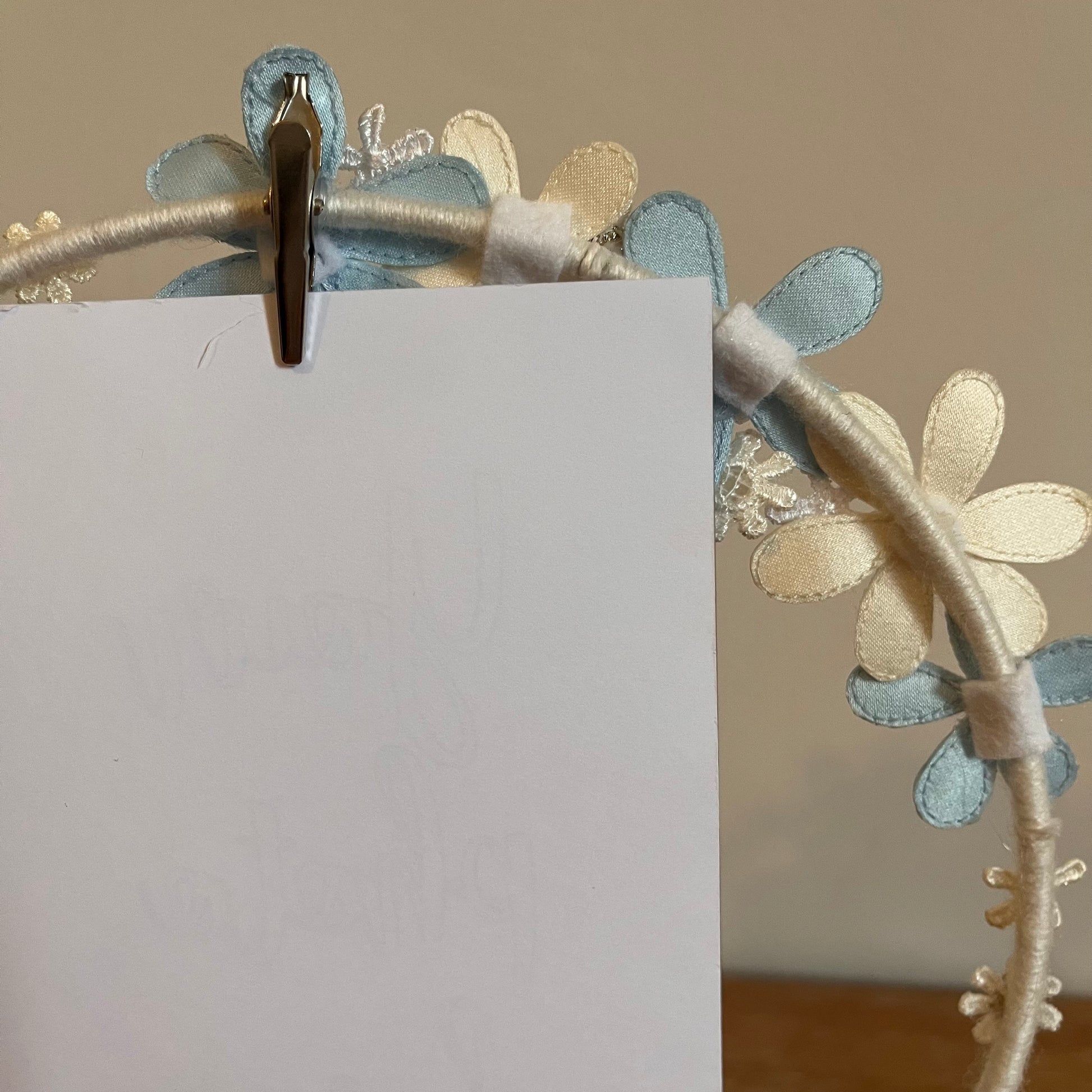 A white card is held by a silver clip on a floral headband with blue and cream fabric flowers.