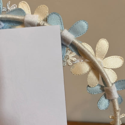Close-up of a headband with light blue and cream fabric flowers, a white card partially obscures the view.