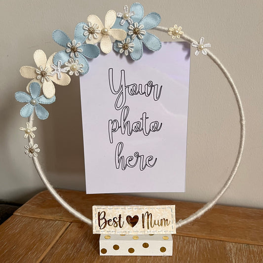 A white hoop photo frame decorated with blue and cream fabric flowers, with a Best Mum sign.