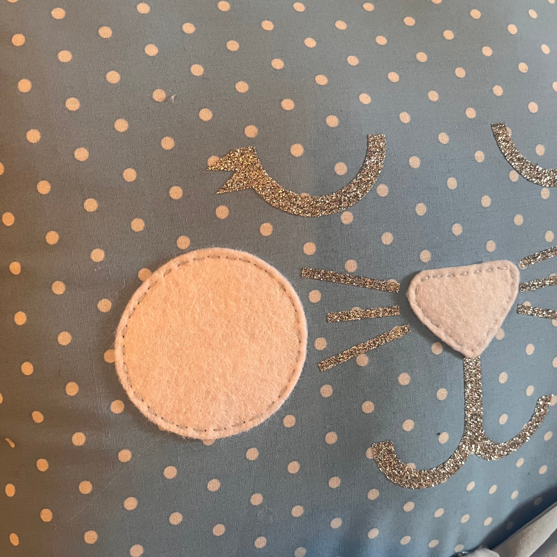 Close-up of a blue polka dot fabric with a cat face design made from felt and glitter.