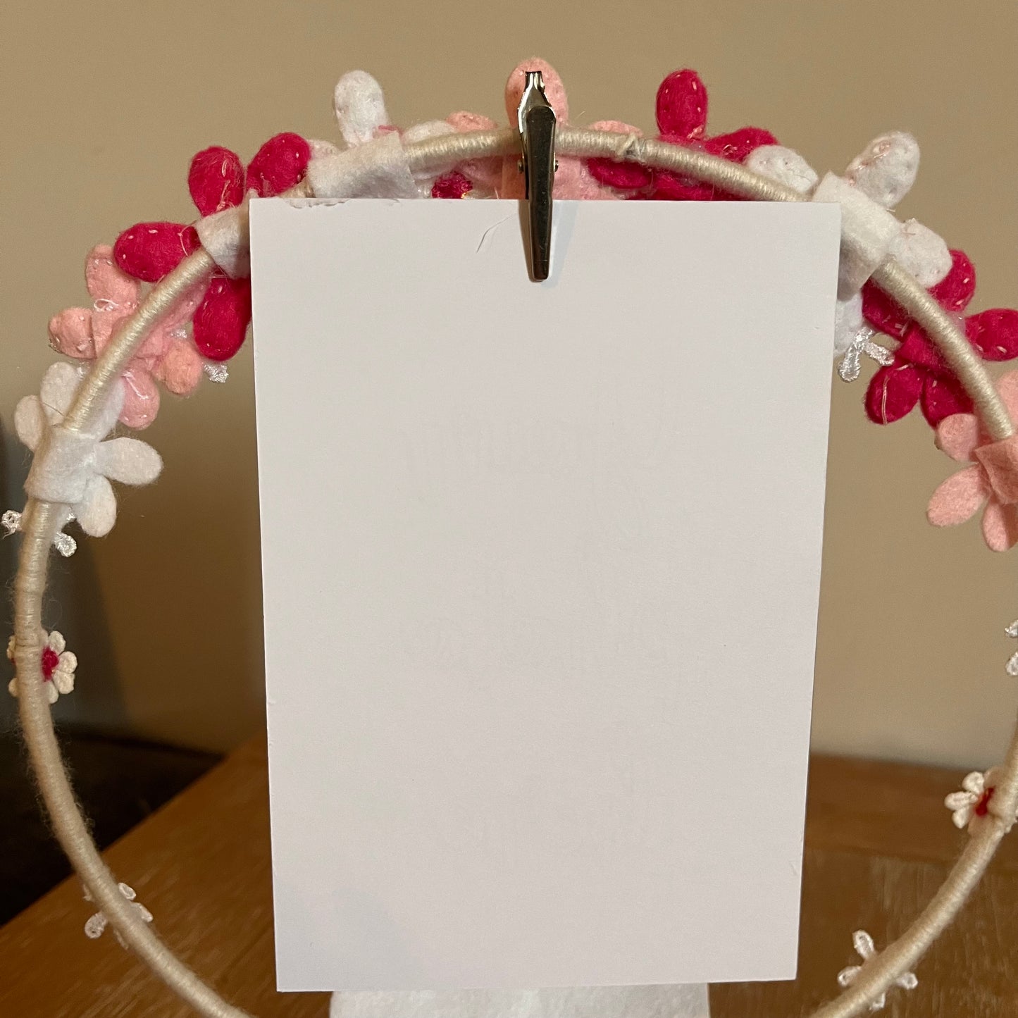 Blank white card held by a silver clip on a floral wreath with pink and white felt flowers.