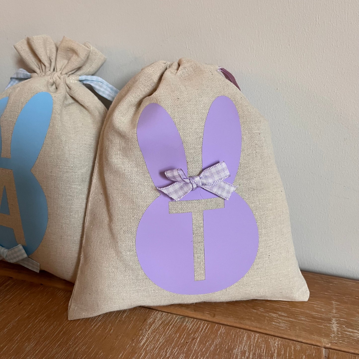 Two linen Easter bunny bags with initial cutouts, one purple T and one blue A, on a wooden surface.