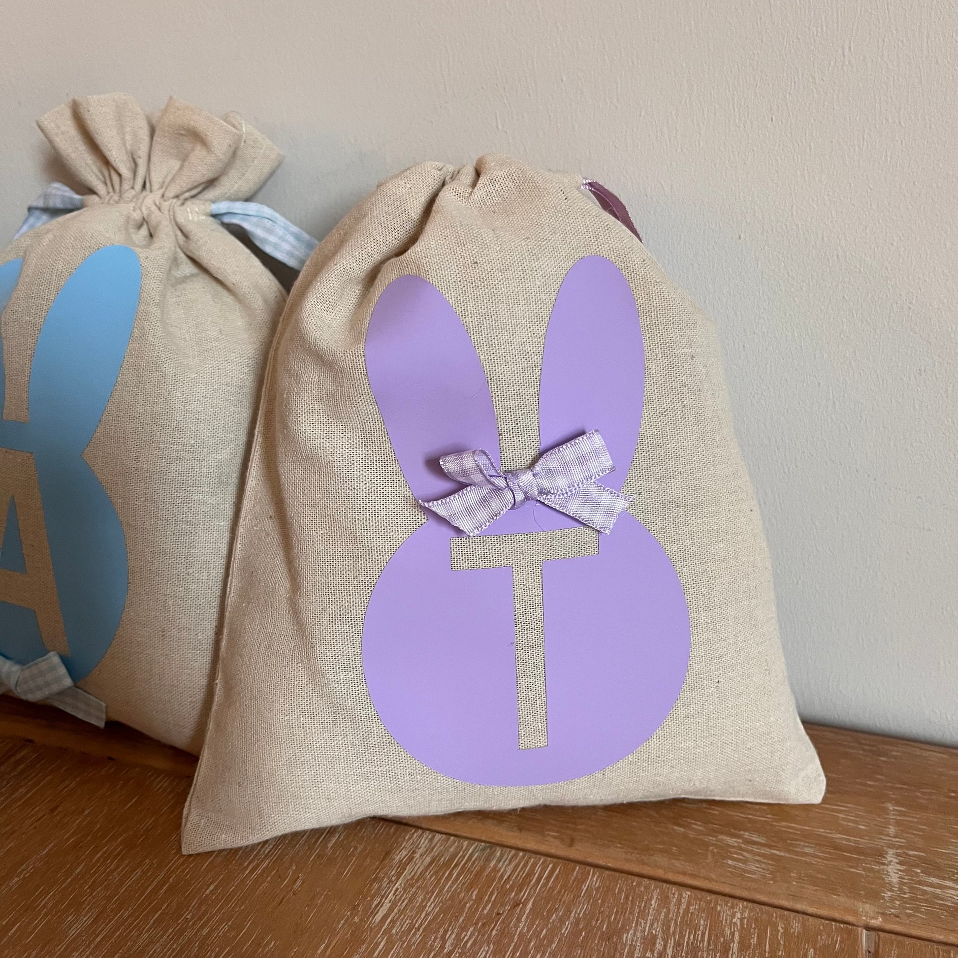 Two linen Easter bunny bags with initial cutouts, one purple T and one blue A, on a wooden surface.