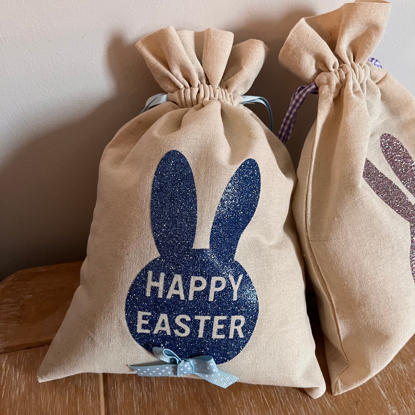 Two linen Easter bags with glitter bunny designs, one blue and one purple, on a wooden surface.