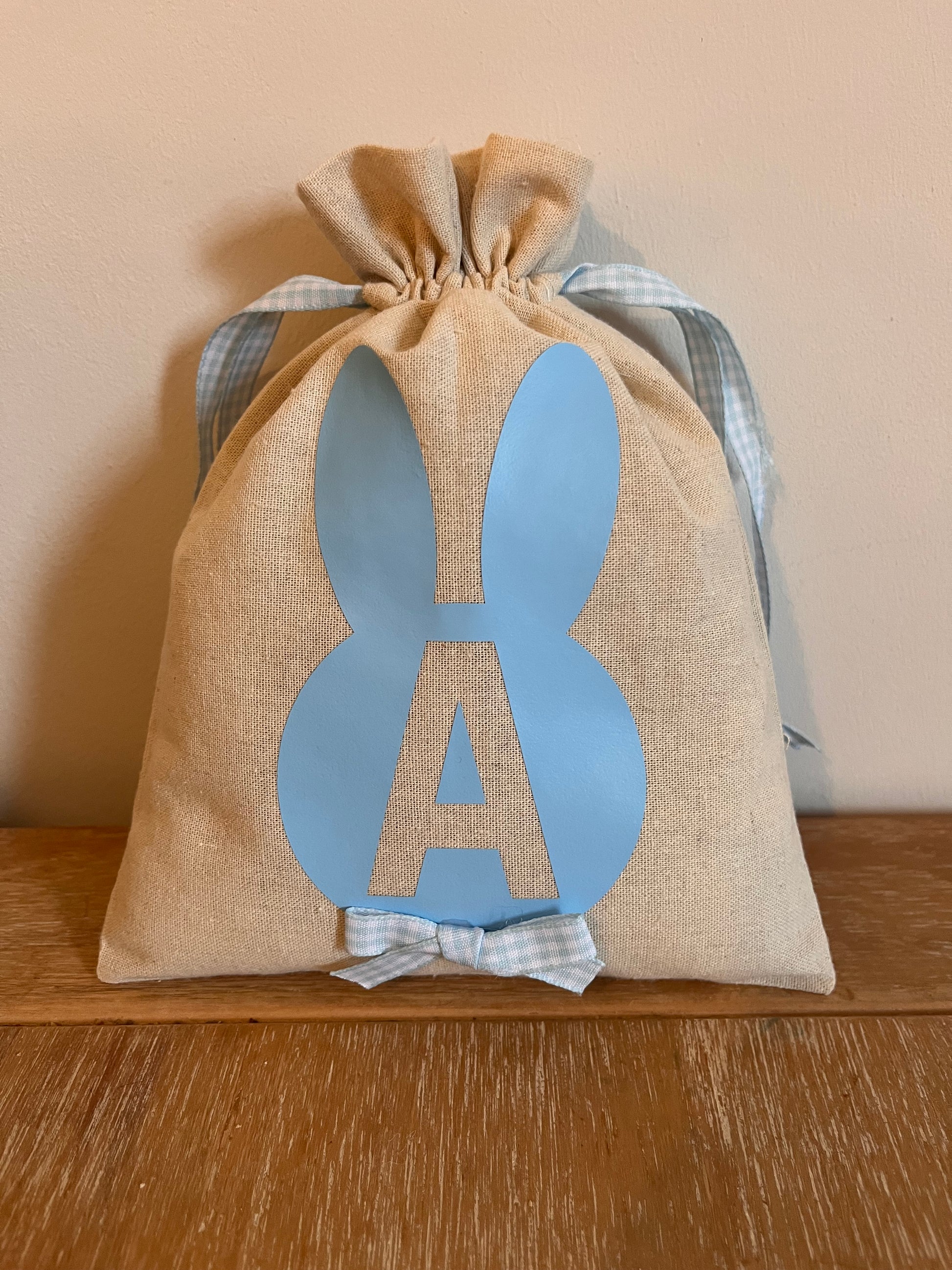 Burlap Easter bunny gift bag with blue ears and a blue letter A, tied with a blue gingham ribbon.