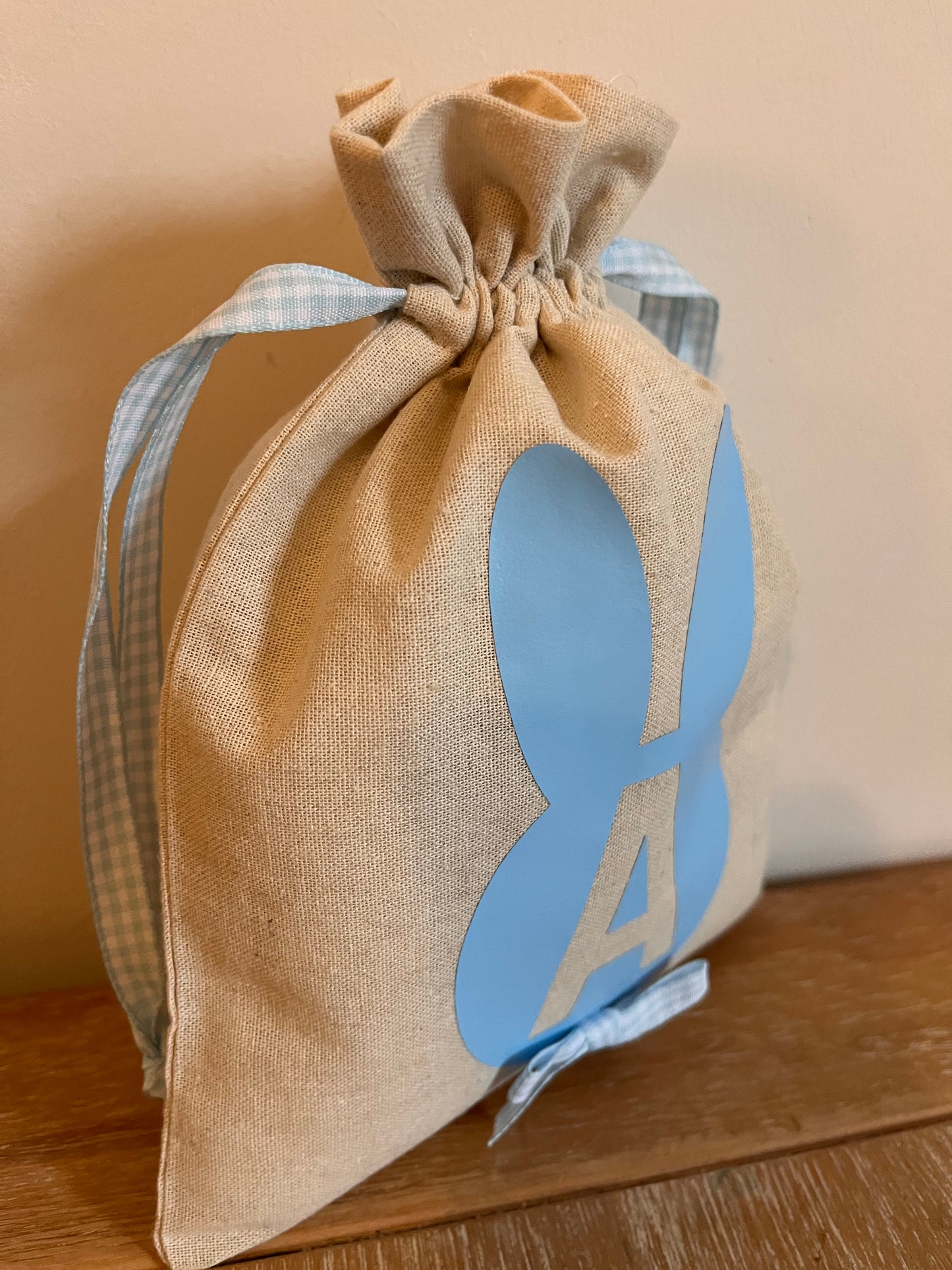 Burlap drawstring bag with blue bunny ears and the letter A, tied with a blue gingham ribbon.