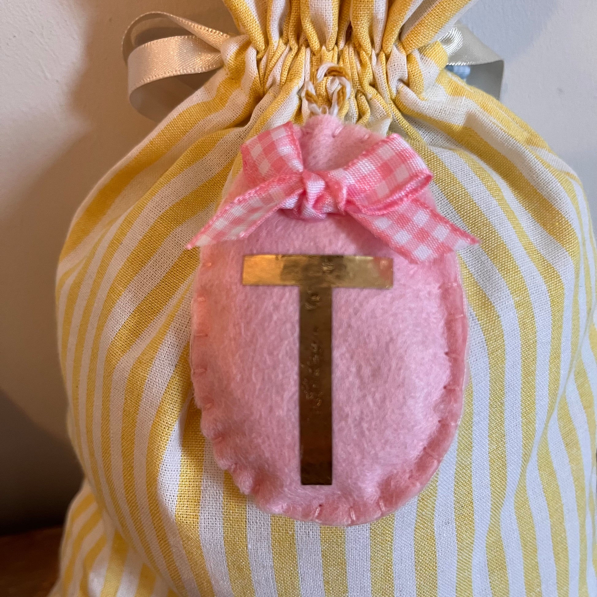 Yellow and white striped drawstring bag with a pink felt egg and a gold letter T.