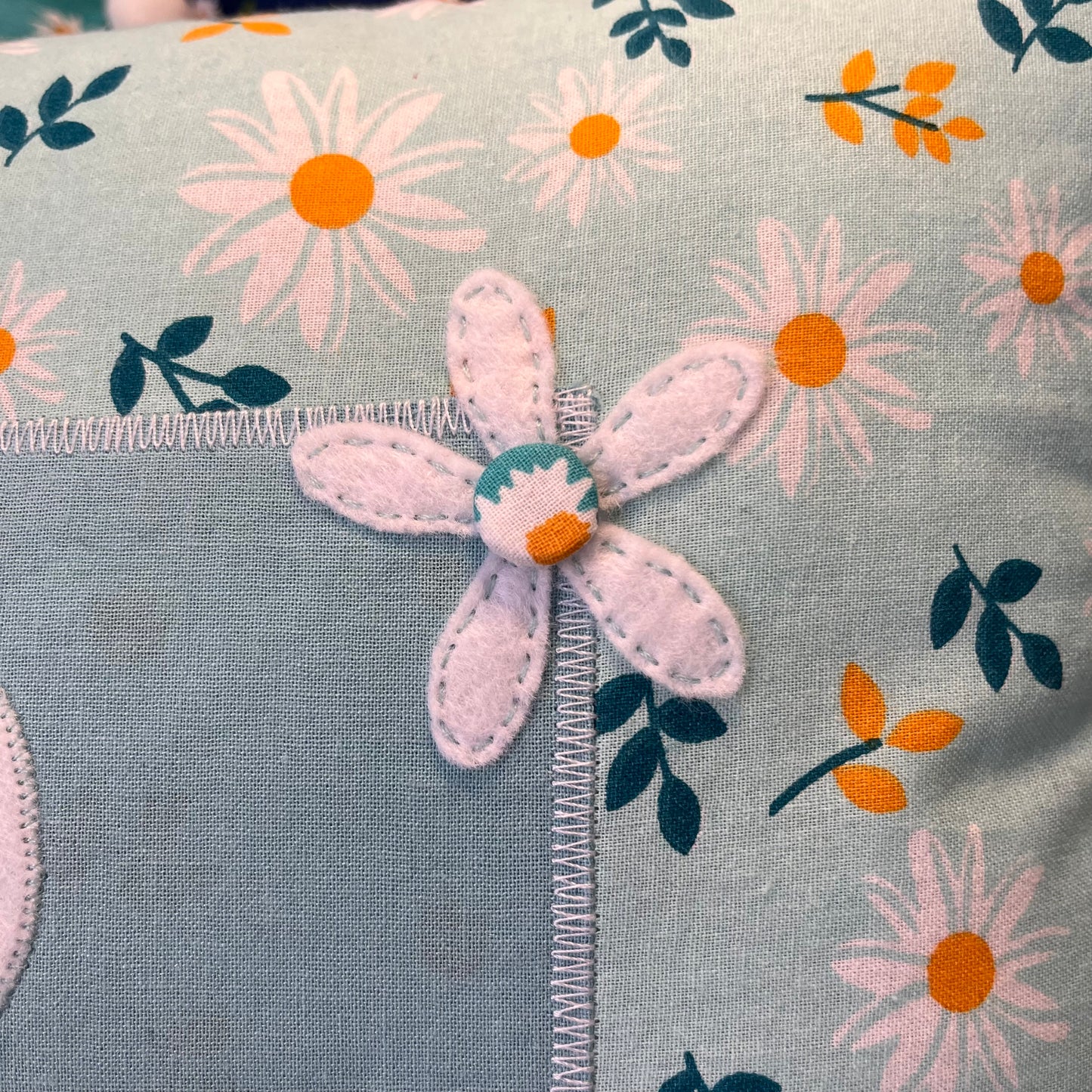 Close up of a light blue fabric with a white felt flower and printed daisies and leaves