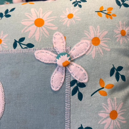 Close up of a light blue fabric with a white felt flower and printed daisies and leaves