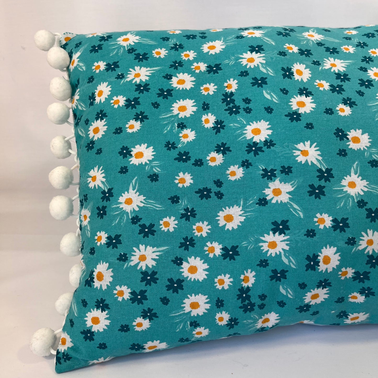 Teal pillow with white and orange daisy pattern and white pom-pom trim