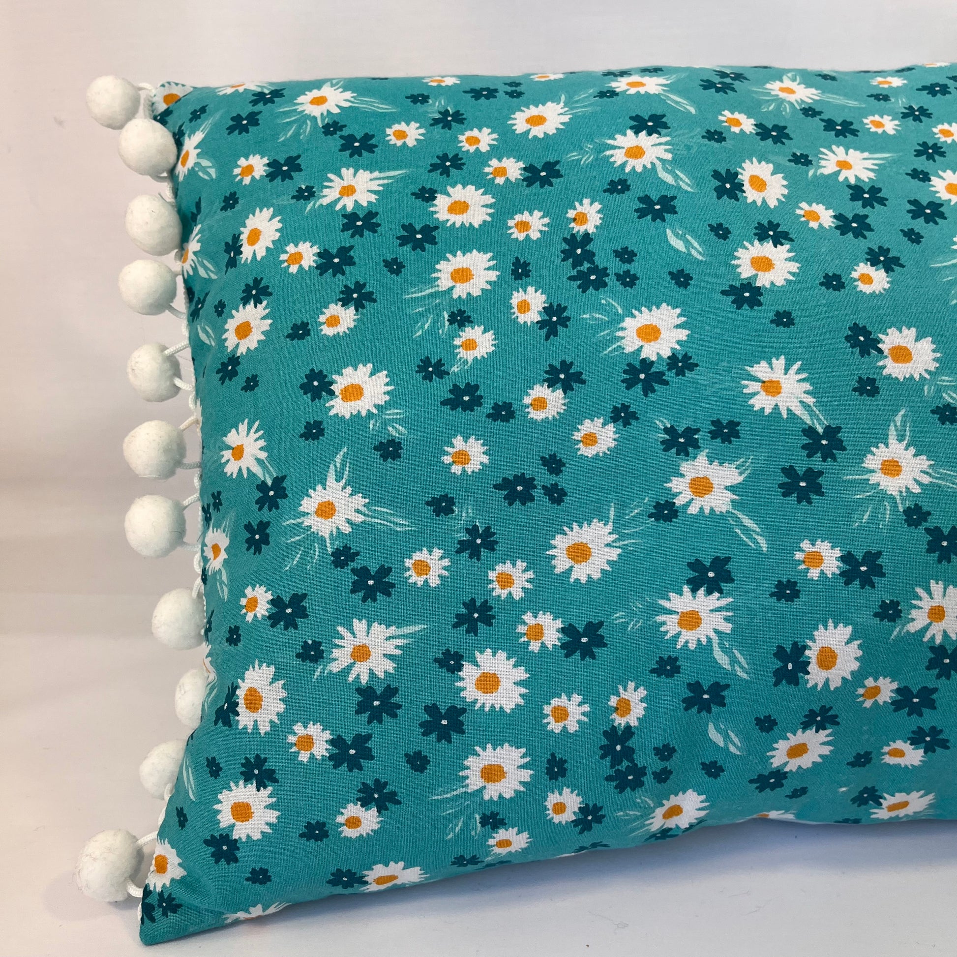 Teal pillow with white and orange daisy pattern and white pom-pom trim