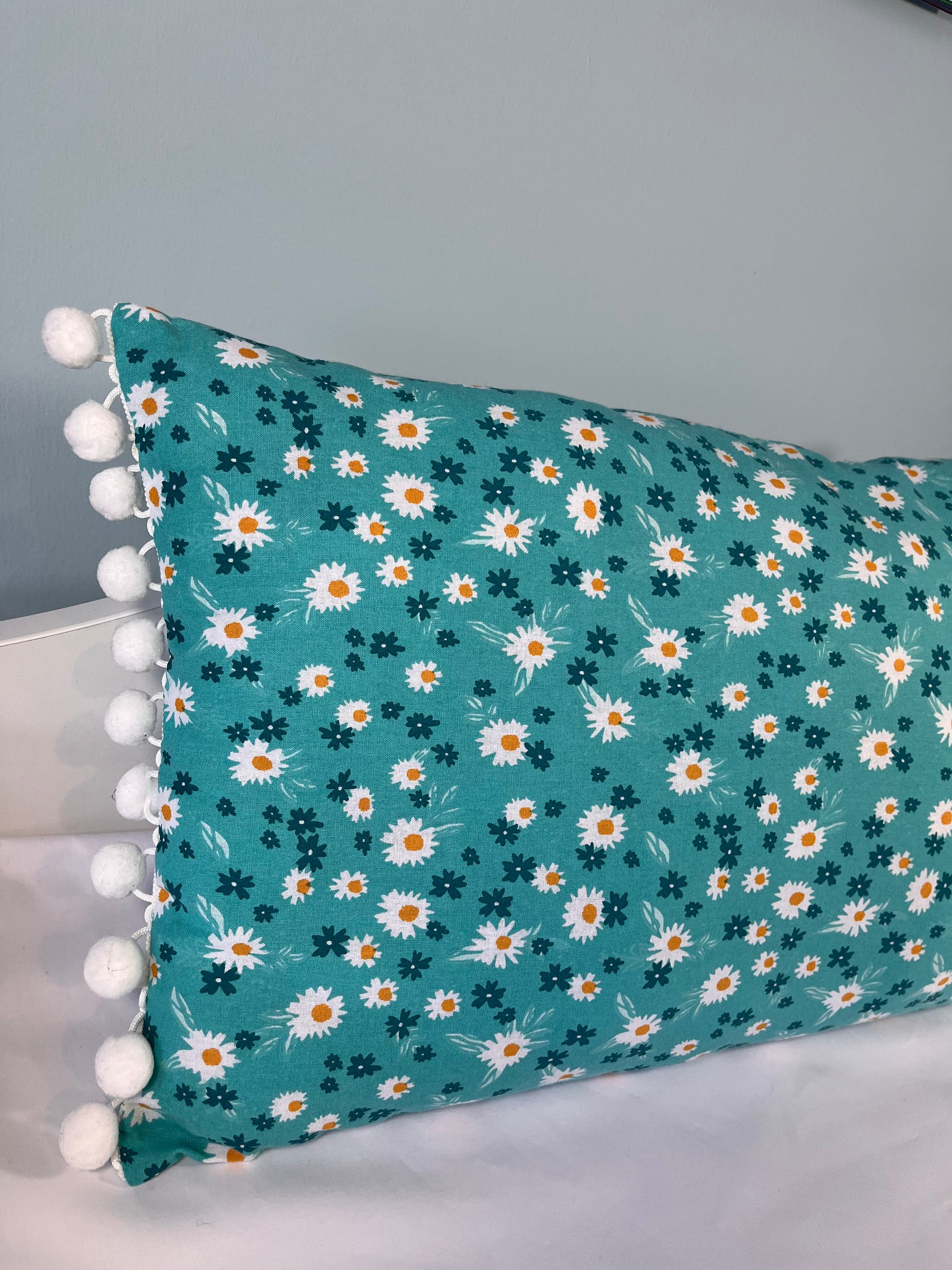 Teal pillow with white and orange daisy pattern and white pom-pom trim