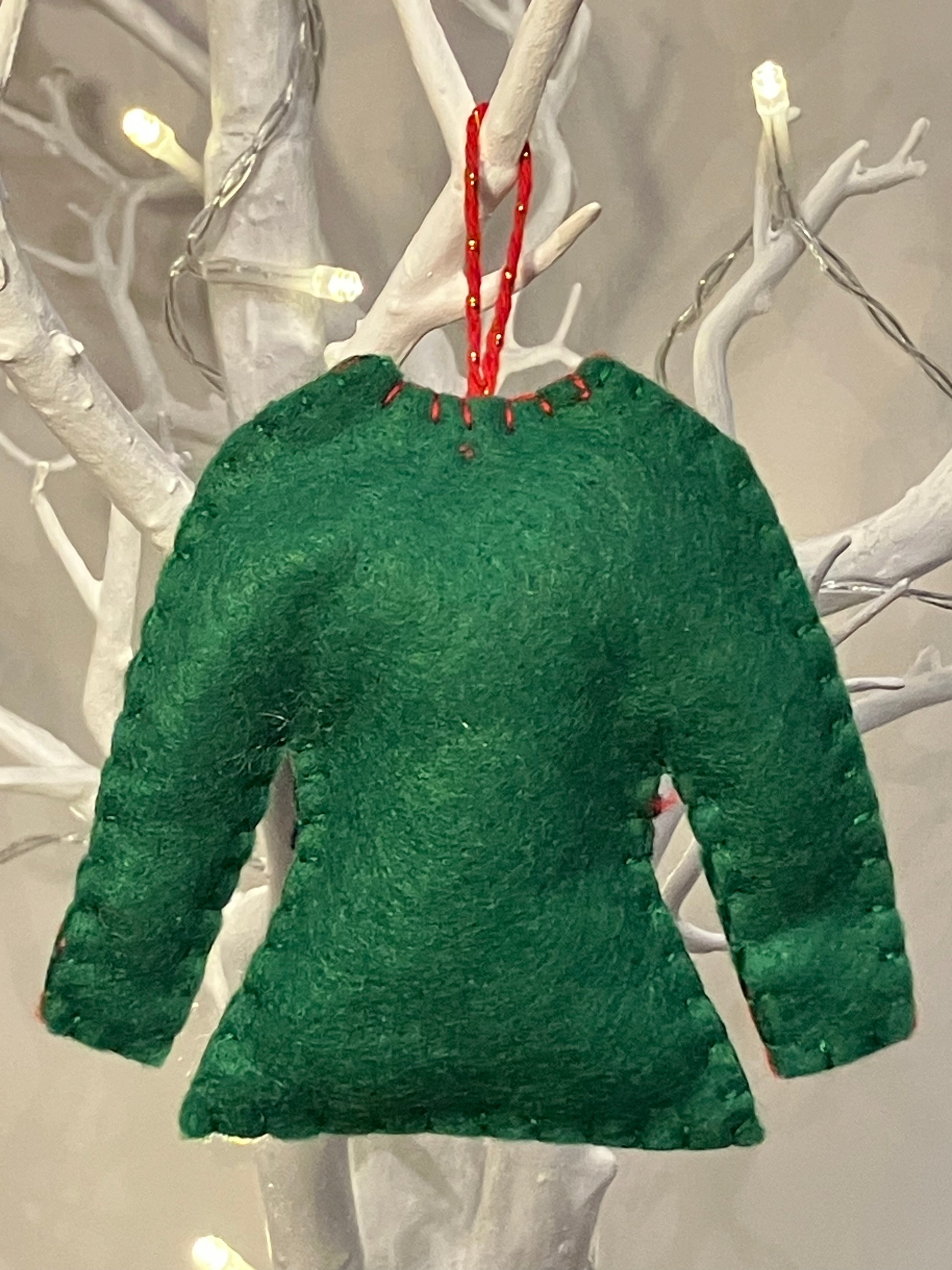 Back of green felt elf shirt ornament with red hanging string on a white branch.