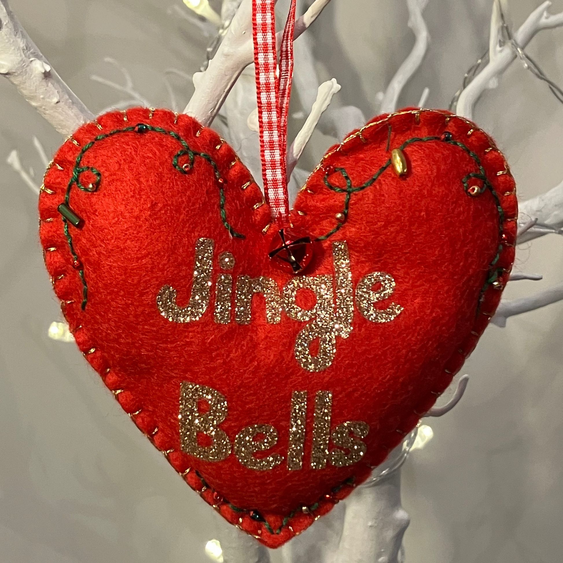 A red felt heart ornament with Jingle Bells in gold glitter, hanging from a branch.
