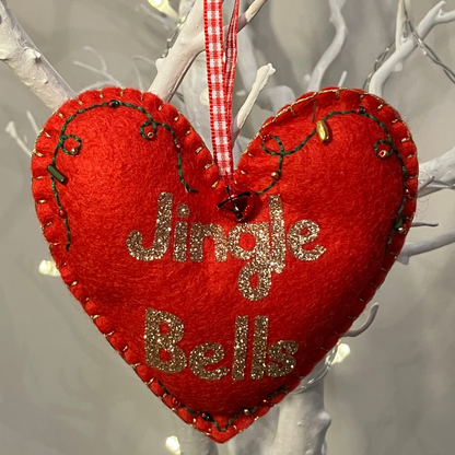 A red felt heart ornament with Jingle Bells in gold glitter, hanging from a branch.