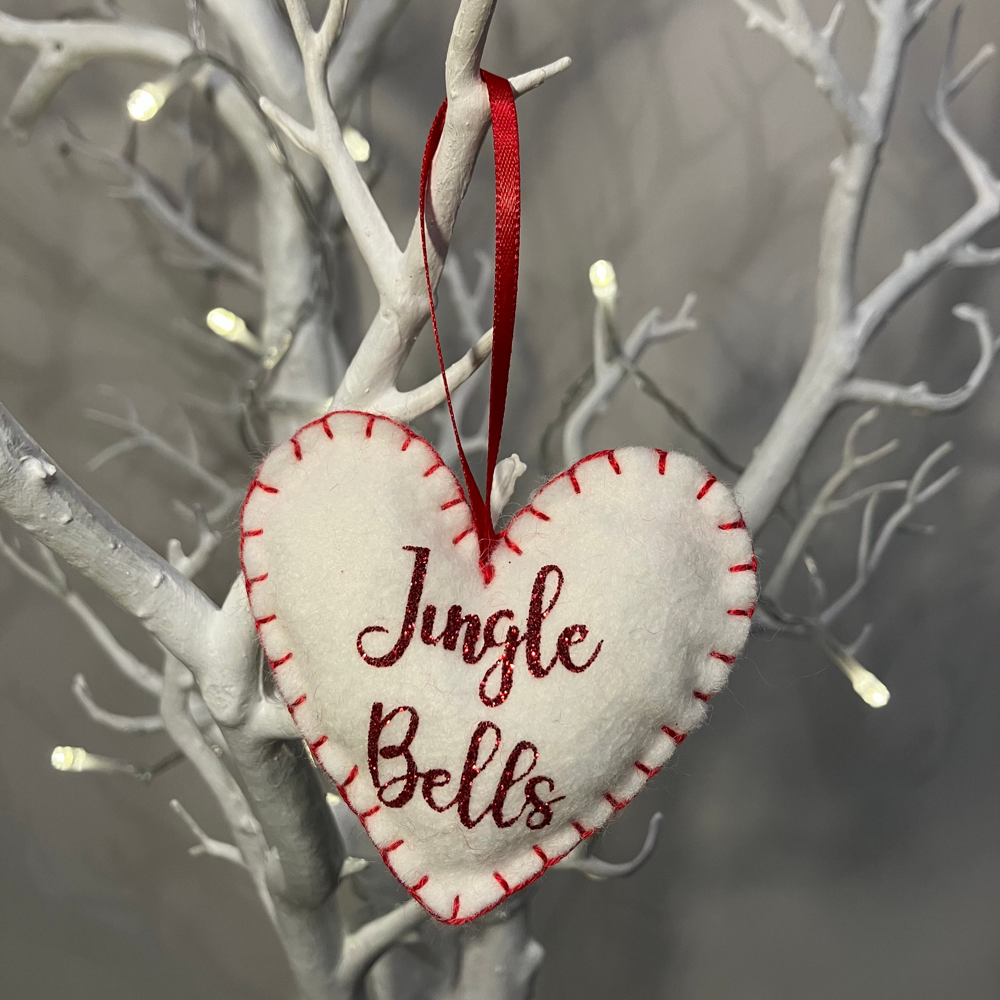A white felt heart ornament with Jingle Bells in red glitter, hanging from a branch.