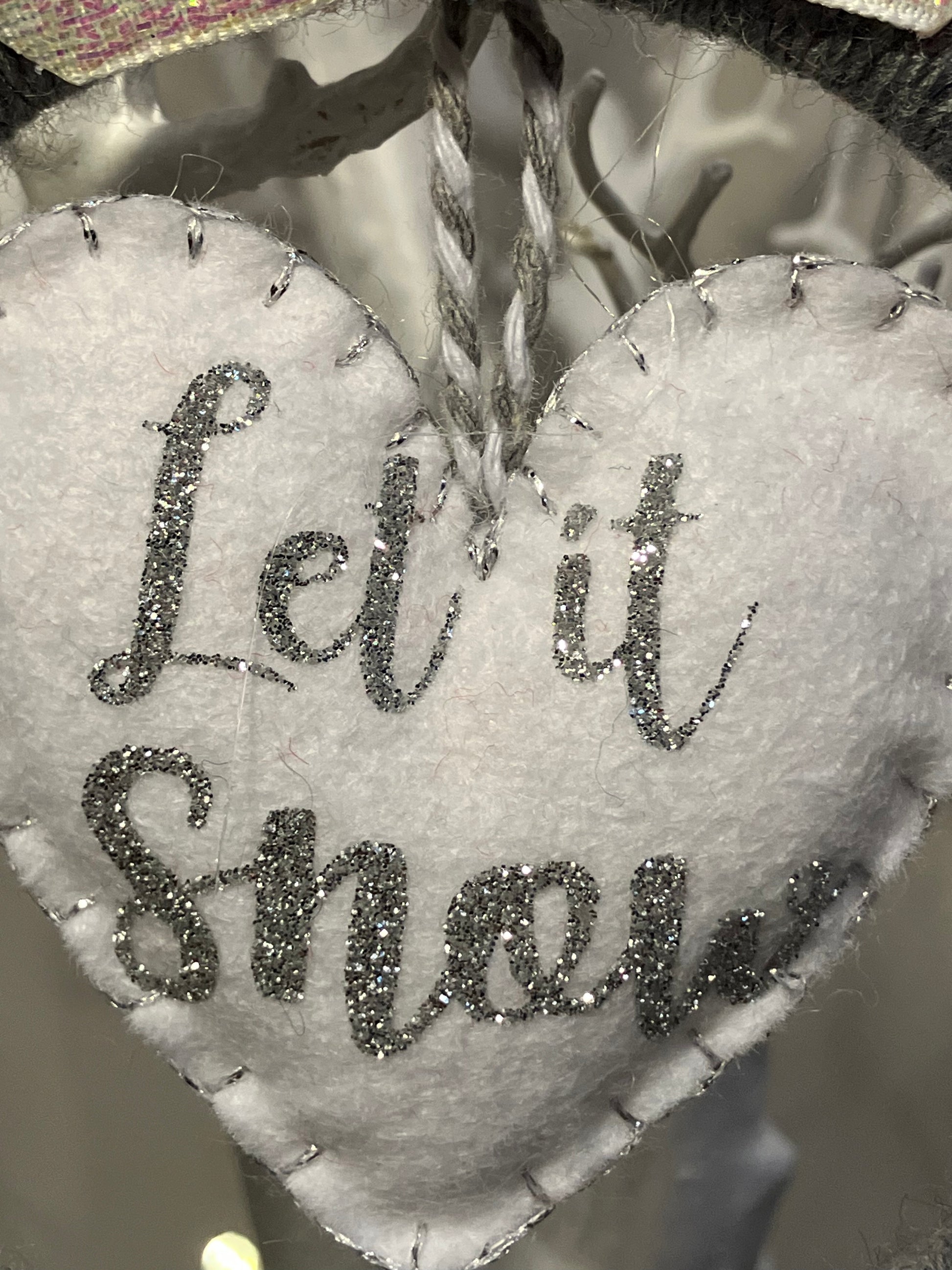 Close-up of the sparkly gray Let it Snow text on a white felt heart ornament.