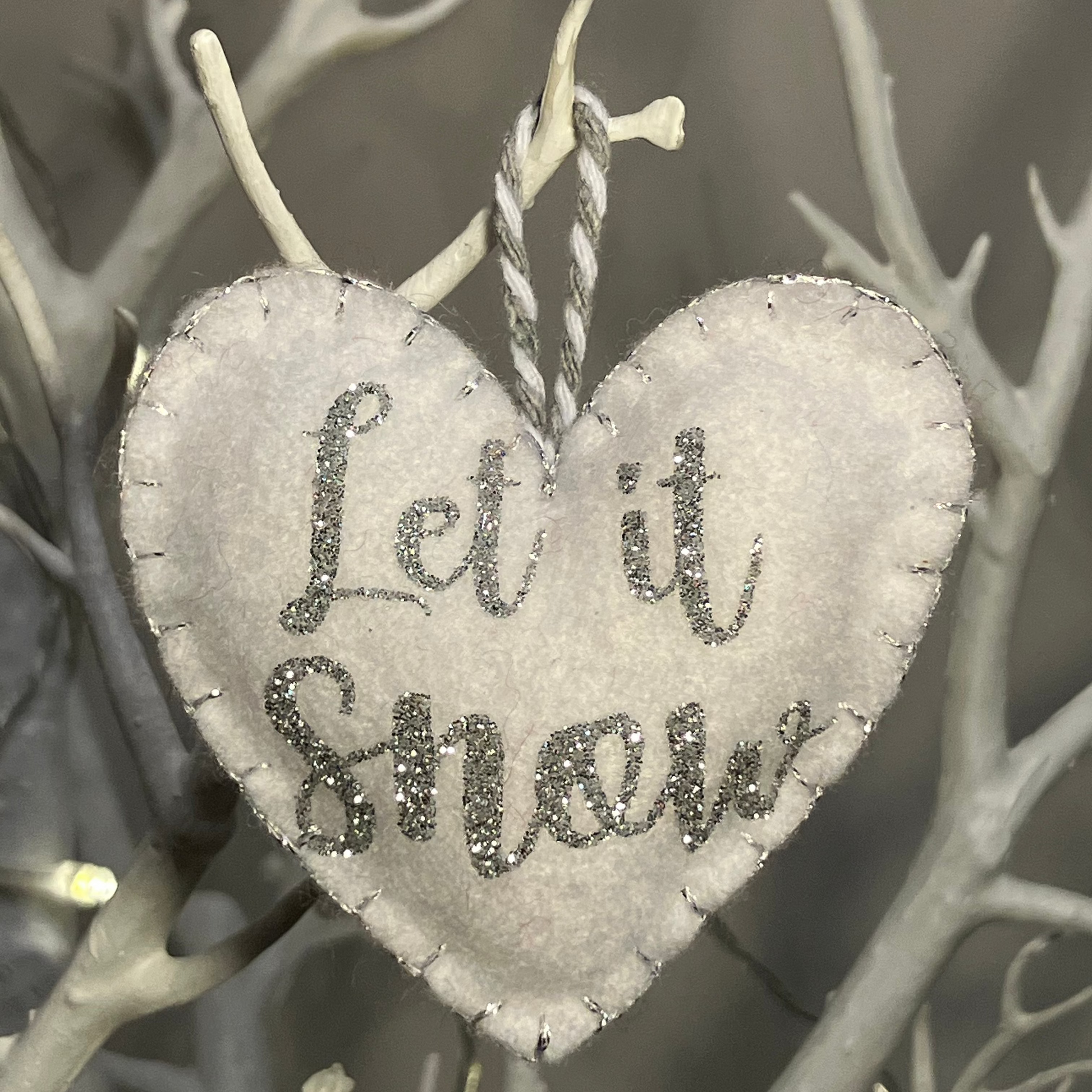 A white felt heart ornament with Let it Snow in silver glitter, hanging.