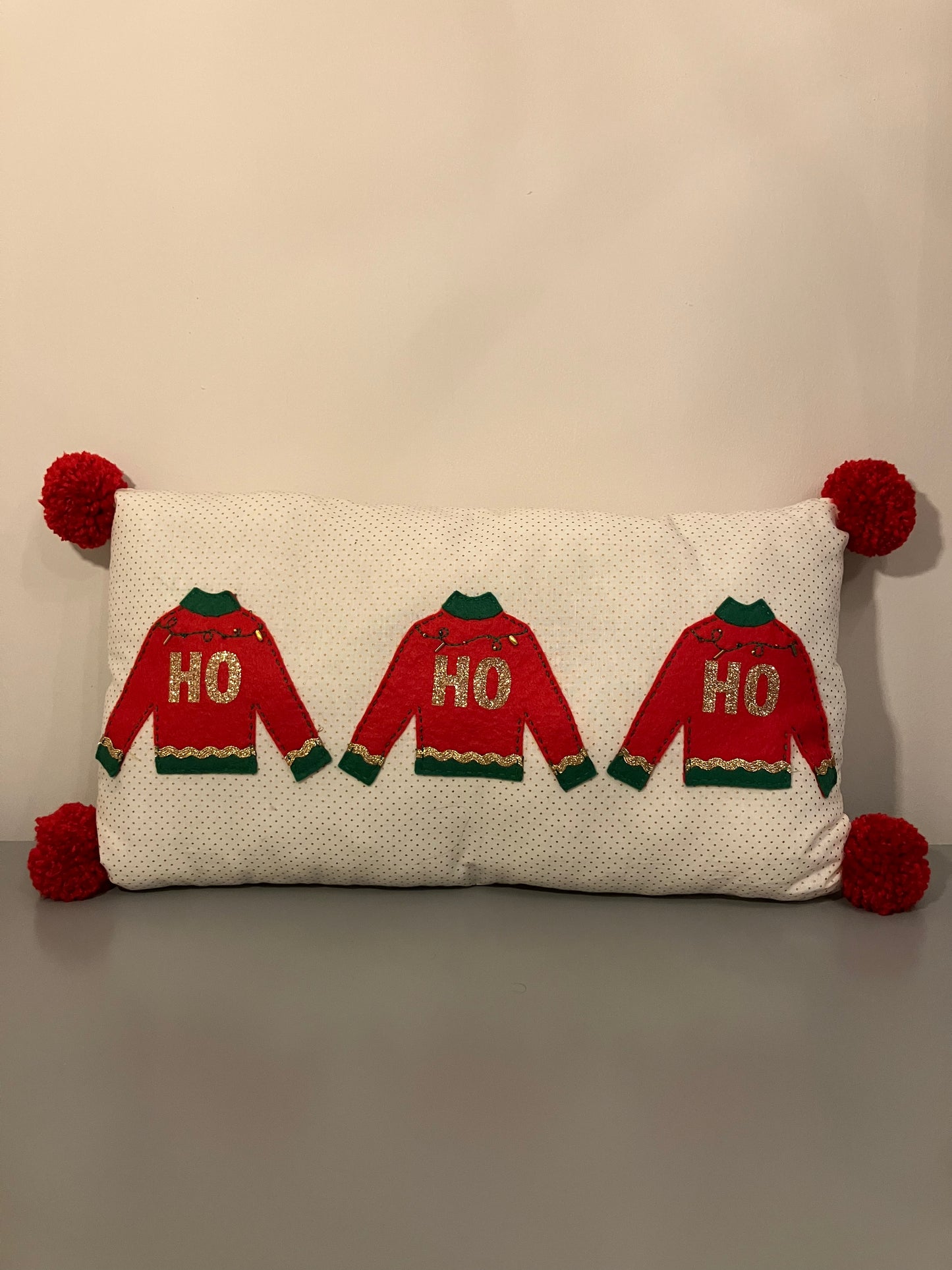 A festive pillow with three red "HO HO HO" sweaters and red pom-poms on the corners.
