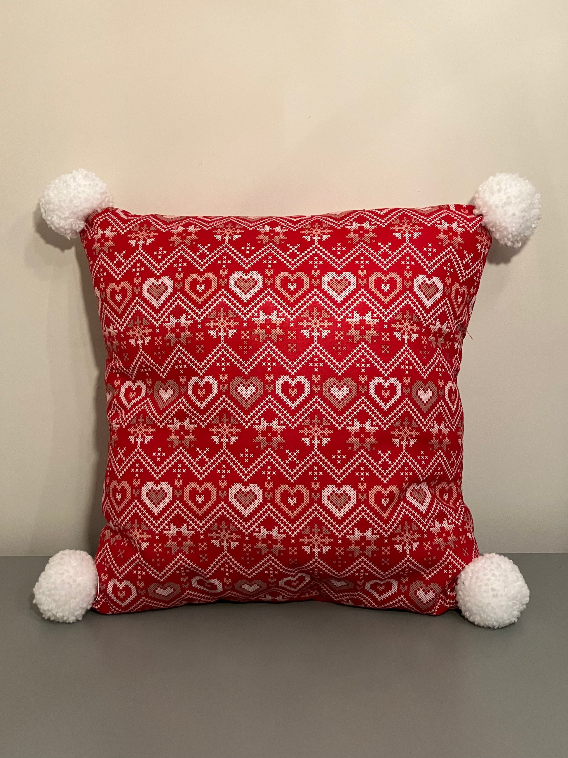 A red pillow with a white and gold heart pattern, with white pom-poms on each corner.