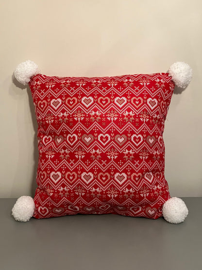 A red pillow with a white and gold heart pattern, with white pom-poms on each corner.