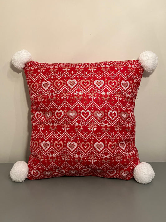 A red pillow with a white and gold heart pattern, with white pom-poms on each corner.