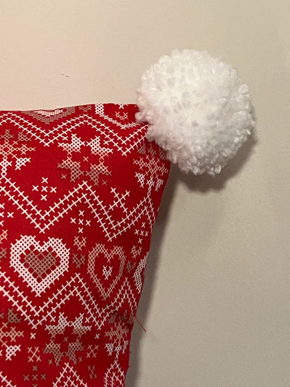 A close-up of a red pillow with a heart pattern and a white pom-pom on the corner.