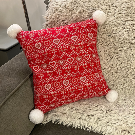 A red pillow with a white and gold heart pattern, resting on a white fuzzy blanket.