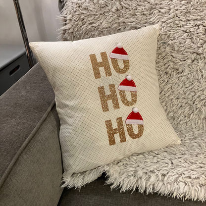 A white decorative pillow with glittery gold 'Ho Ho Ho' text and mini Santa hats.