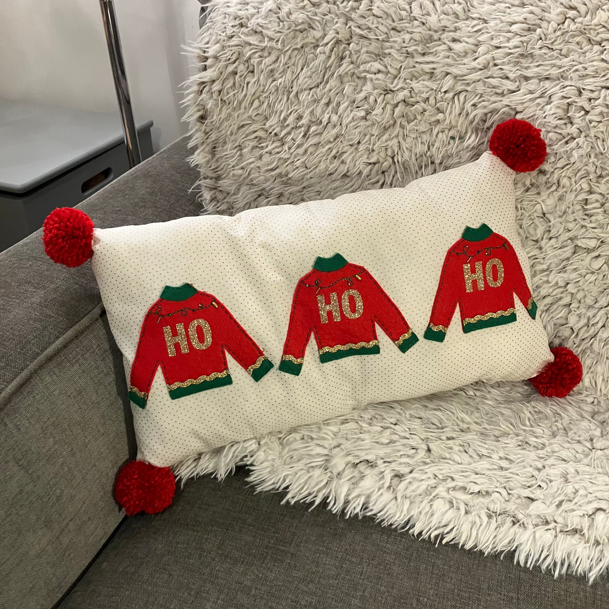 A festive pillow with three "HO HO HO" sweaters and red pom-poms on a grey couch.