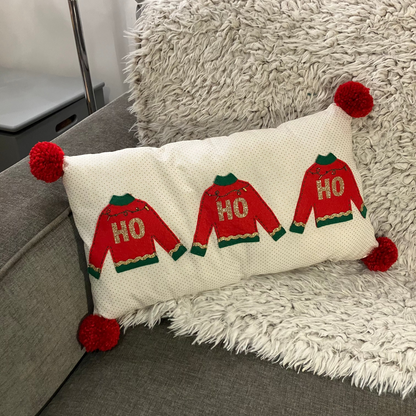 A festive pillow with three "HO HO HO" sweaters and red pom-poms on a grey couch.