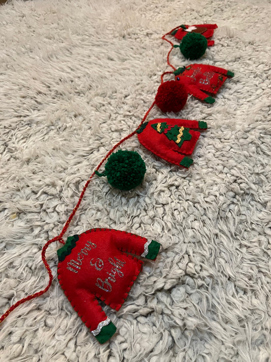 A garland of felt red Christmas sweaters and green pom-poms on a fluffy white surface.