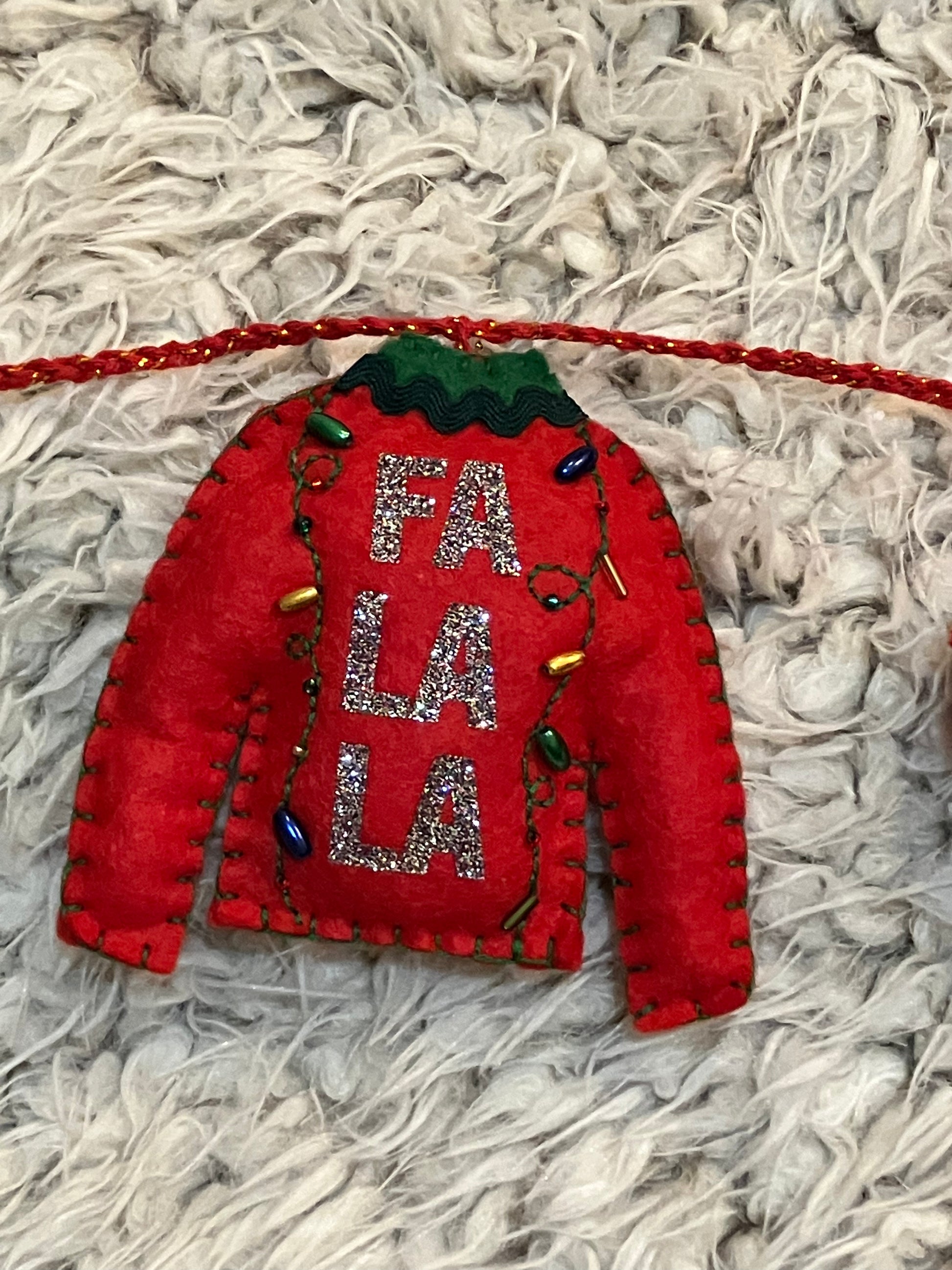 A felt red sweater ornament with "FA LA LA" and tiny Christmas lights on a white surface.