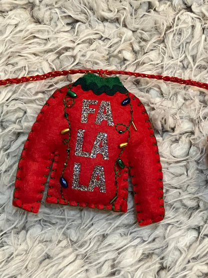 A felt red sweater ornament with "FA LA LA" and tiny Christmas lights on a white surface.
