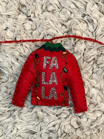 A felt red sweater ornament with "FA LA LA" and tiny Christmas lights on a white surface.