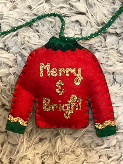 Red felt Christmas sweater ornament with "Merry & Bright" in gold glitter on a fuzzy gray surface.